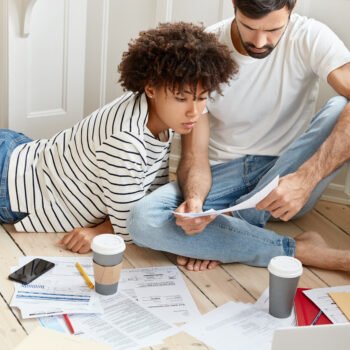 Family couple maintains home accounting, discuss plans and future payments, surrounded with bills on floor, dressed in domestic clothes, drinks tasty hot coffee, analyze income chart together